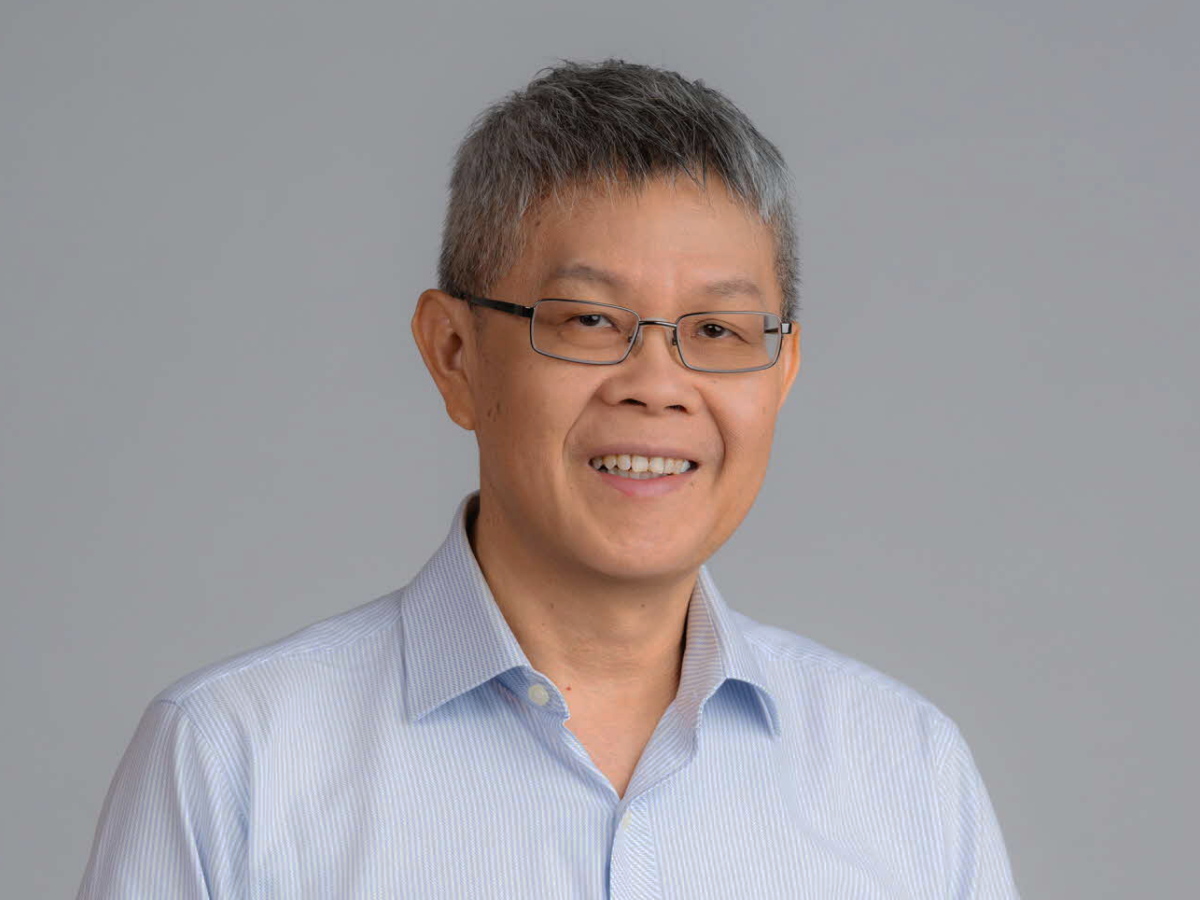 Mr Lim Boon Wee in a light blue shirt smiling with hands clasped and resting on a chair against a plain grey background